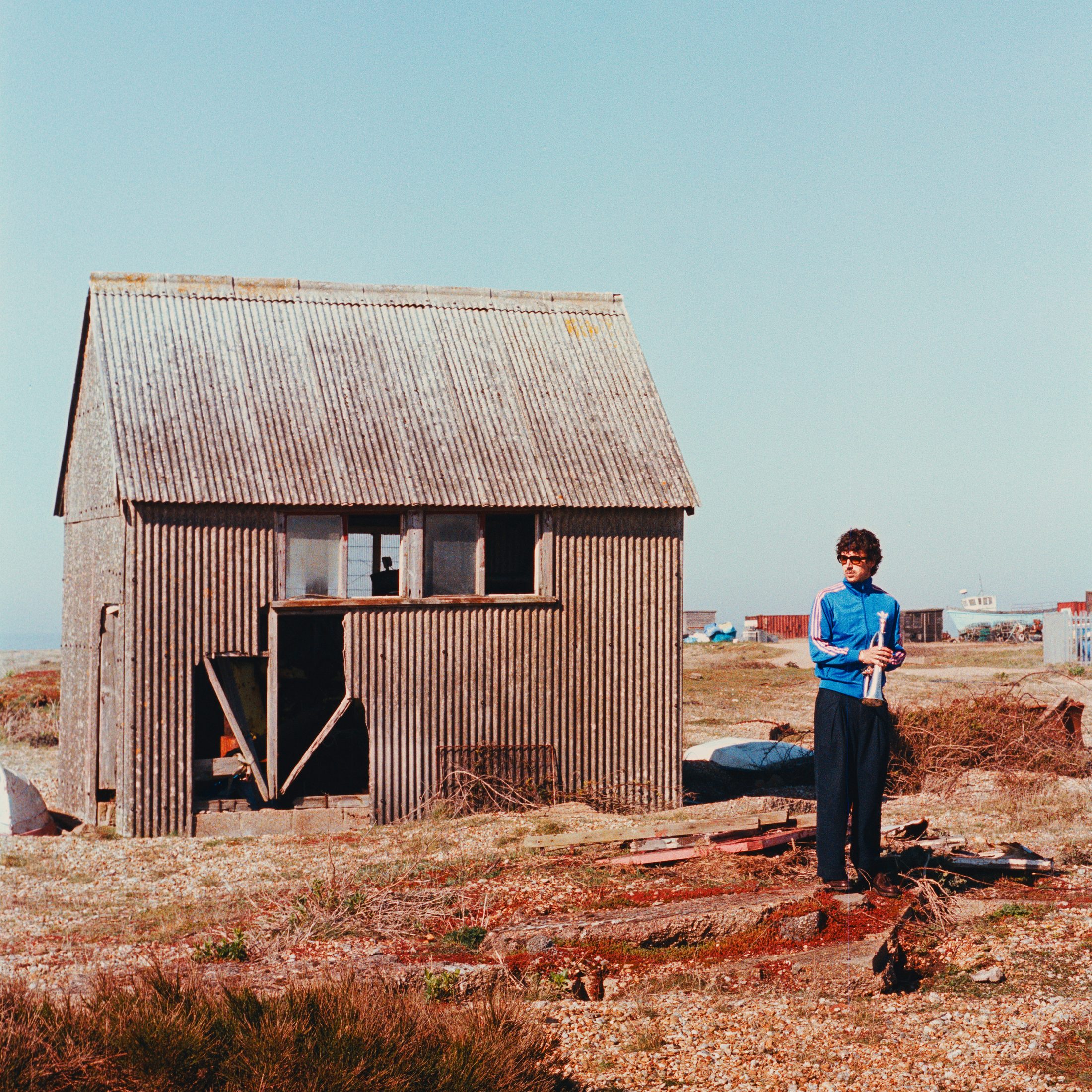 Sol Paradise standing beside a weathered shed on a shingle beach, holding a trumpet